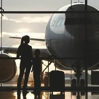 Silhouettes of a woman and child with luggage looking at an airplane through airport windows at sunset.